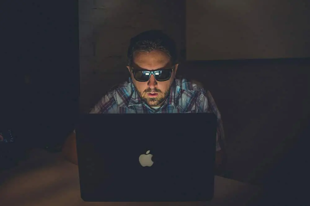 Man wearing sun glasses while sitting in a dark room in front of a laptop