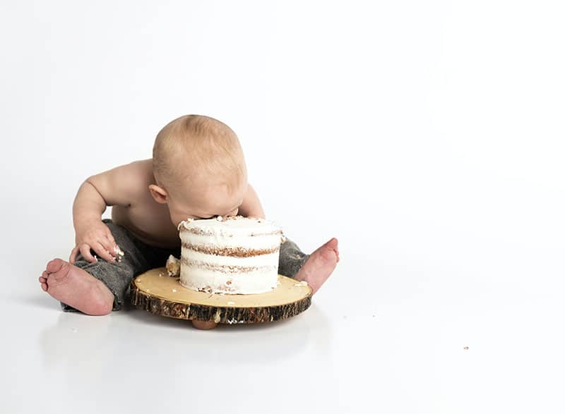 A cake with white icing on wood with a baby sitting with his legs around it and his face in the cake
